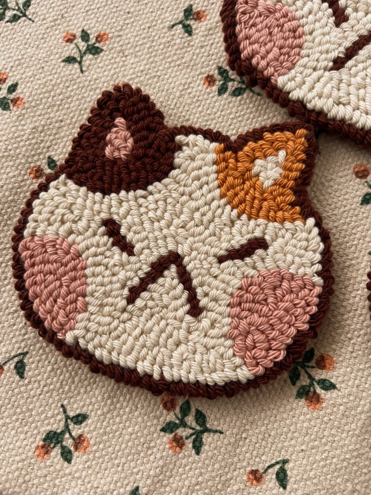It’s a cat head made of cotton that can be used to decorate or place its cup/glass. The cat is beige, with big pink cheeks and an orange ear and a brown ear. It’s a punch needle coaster.