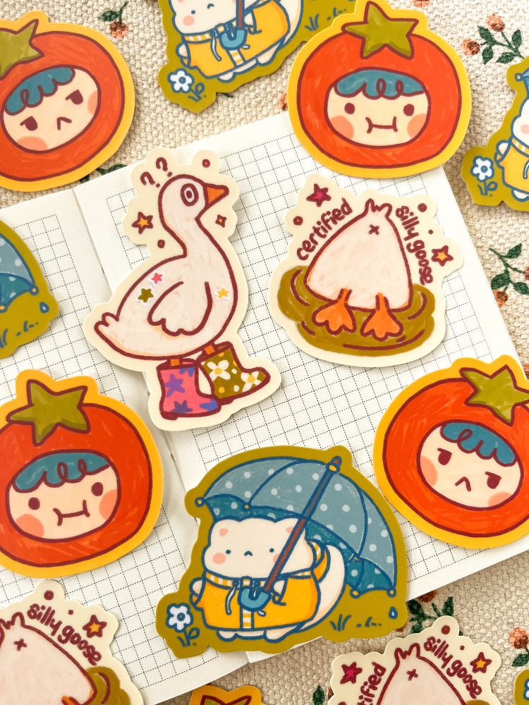 Collection of colorful stickers featuring cute characters: a goose in boots, a goose with his head in the water, a tomato-head figure, and Yume the cat with an umbrella, arranged on graph paper.