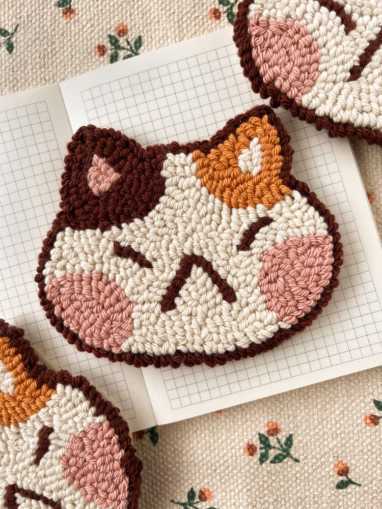 It’s a cat head made of cotton that can be used to decorate or place its cup/glass. The cat is beige, with big pink cheeks and an orange ear and a brown ear. It’s a punch needle coaster.