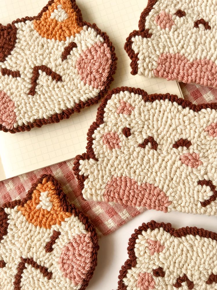 Five handmade punch needle cat coasters on a checkered fabric and notebook background