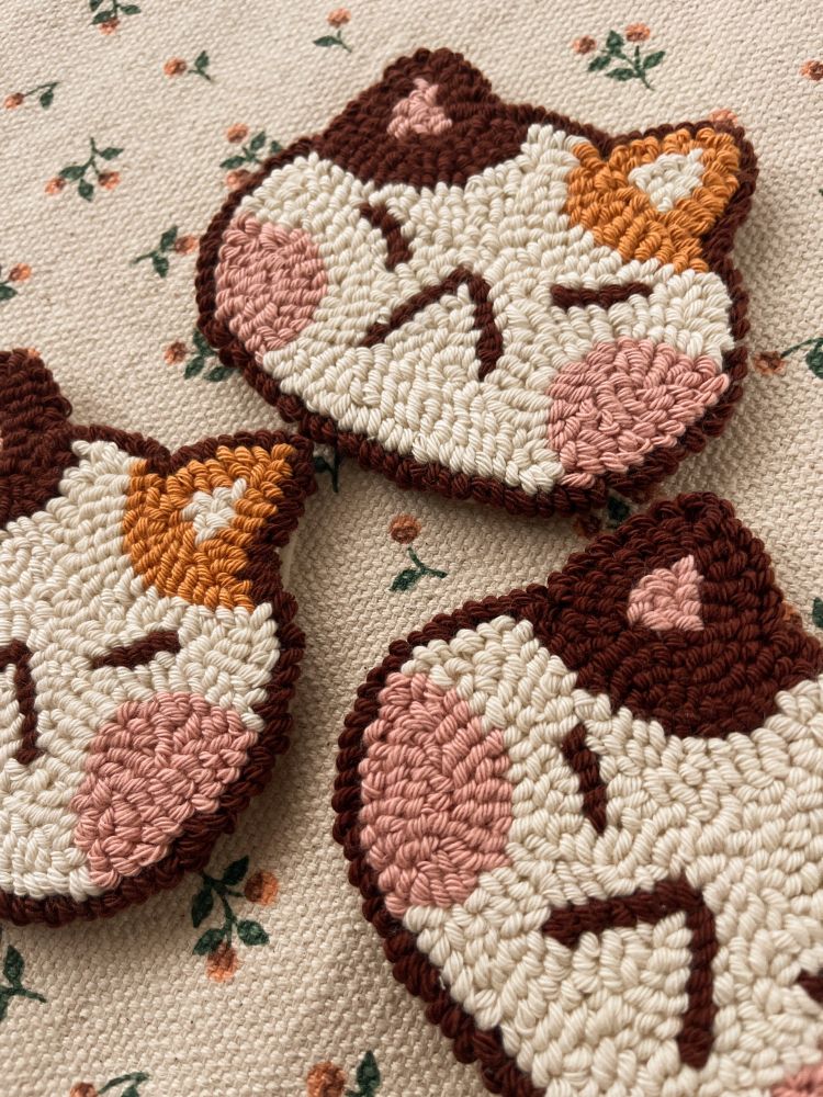 It’s a cat head made of cotton that can be used to decorate or place its cup/glass. The cat is beige, with big pink cheeks and an orange ear and a brown ear. It’s a punch needle coaster.