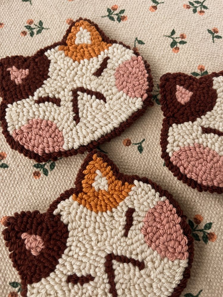 It’s a cat head made of cotton that can be used to decorate or place its cup/glass. The cat is beige, with big pink cheeks and an orange ear and a brown ear. It’s a punch needle coaster.
