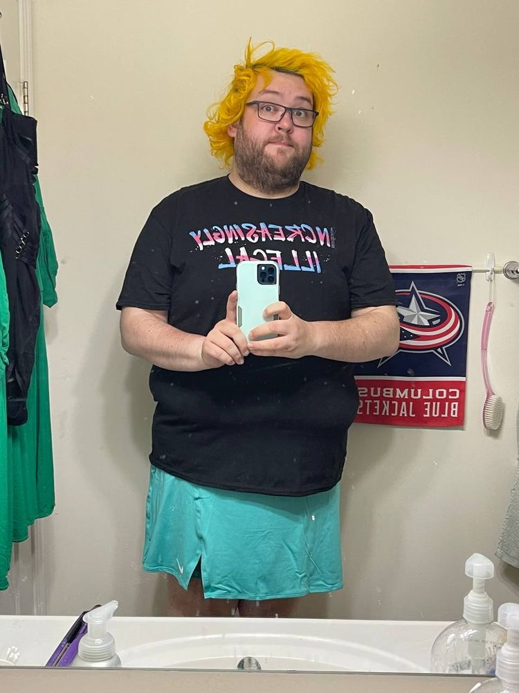 Maddie with yellow hair wearing a teal tennis skirt and a shirt that says increasingly illegal in trans colors