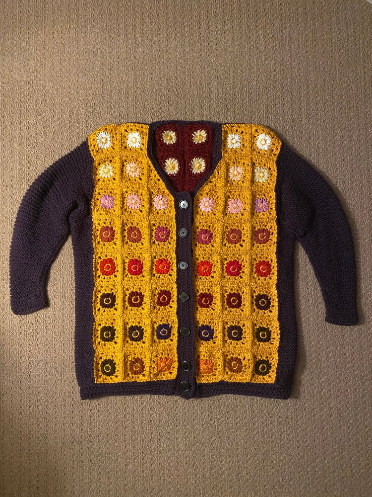 Colour photo: a thick cardigan shown from the front. The front panels are made of vintage daisy maker daisies in gold granny squares, fading from white to brown. The sleeves and side panels are knit in purple, with a crochet button band of the same colour.