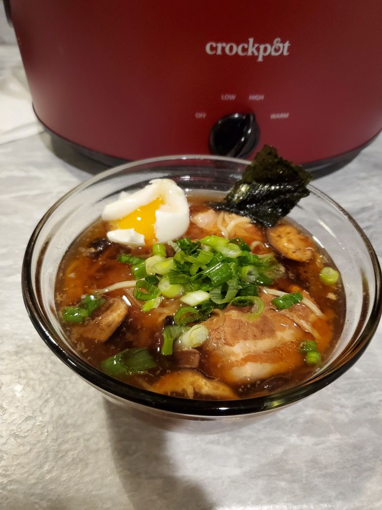 A glass bowl filled with pork ramen in front of a red crockpot. The ramen is topped with seaweed, green onions, and a soft-boiled egg.