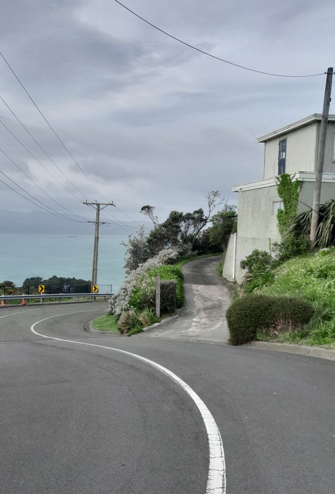 A steep road curves down and a driveway curves up to two-storey building. On the second story, a blue door with window panes gives out onto the roof of the first storey. There's no balustrade or railing. In the background is the sea and distant misty hills.
