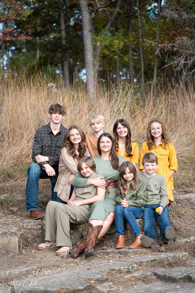 A portrait of two mothers (sisters) and their children, all sitting on a set of low, rocky steps outside. The mother in center, wearing a sage green dress, sits in the middle with a child on her lap and another leaning on her. Behind her to her left is the second mother, dressed in mustard yellow. The children, a mix of boys and girls of various ages, are wearing coordinated fall-toned clothing, including plaid, yellow, tan, and olive green. They are backed by tall brown grass and a wooded area.
