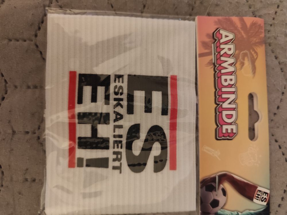 Eine ovp Armbinde in weiß, auf der  ES ESKALIERT EH! steht. Oben und unten durch eine rote Linie eingerahmt. 