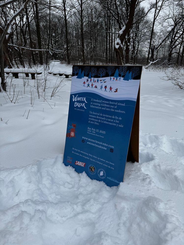 An a-frame sign set up in a snowy forest describing the schedule of winter break MKE, a winter festival at havenwoods state park