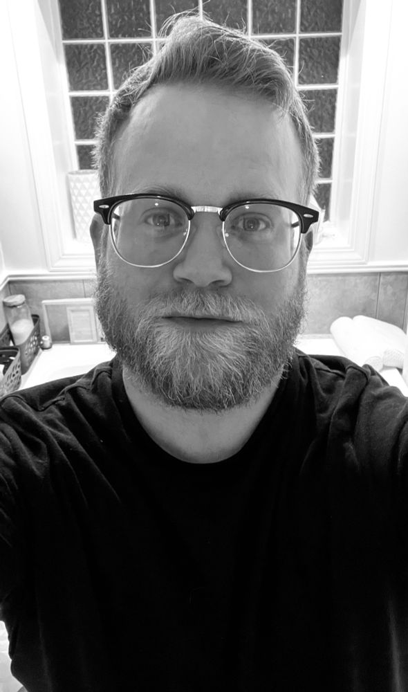 A black and white photo of me wearing glasses. Blonde hair blue eyed man (although you can’t tell eye color here) wearing a dark shirt and giving a slight grin to the camera 