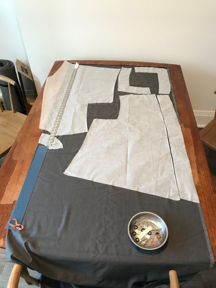 A vest pattern laid out on grey cloth on a wooden table, with rulers and a metal pin bowl holding the ends down. The pattern the Ashland vest is by Diane Ericson.