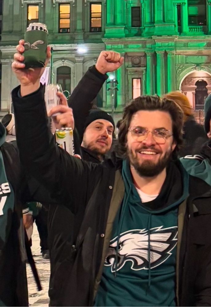 Man in midnight green Eagles sweatshirt standing on North Broad Street, smiling and raising his right hand while holding a drink with an old school Eagles logo koozie, with Philadelphia City Hall illuminated in green in the background