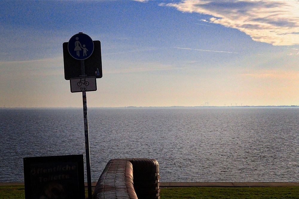 Die Nordsee, ein bisschen Gras, eine Ziegelsteinmauer und ein Schild für einen Fußweg/Radfahrer Frei. Ein Schild "öffentliche Toulette" steht daneben. Blauer Himmel, man sieht ein paar Wolken