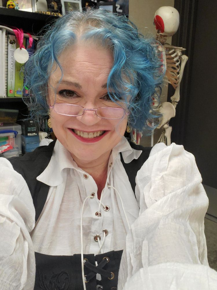 Me, a white woman with curly blue hair is facing the camera and smiling broadly. I'm dressed in a blousy white shirt and black bodice and am wearing small rectangular reading glasses perched on my nose. Behind me is a bookshelf and laboratory skeleton. 