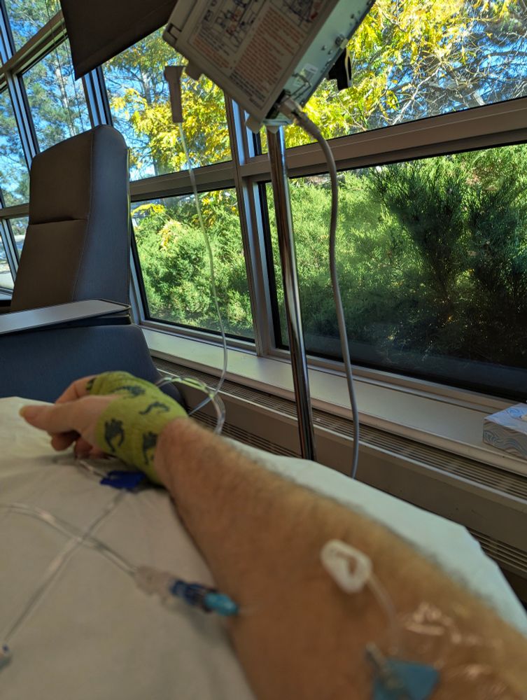 White man's arm with chemo drugs being infused. Beautiful greenery seen through the windows in background 