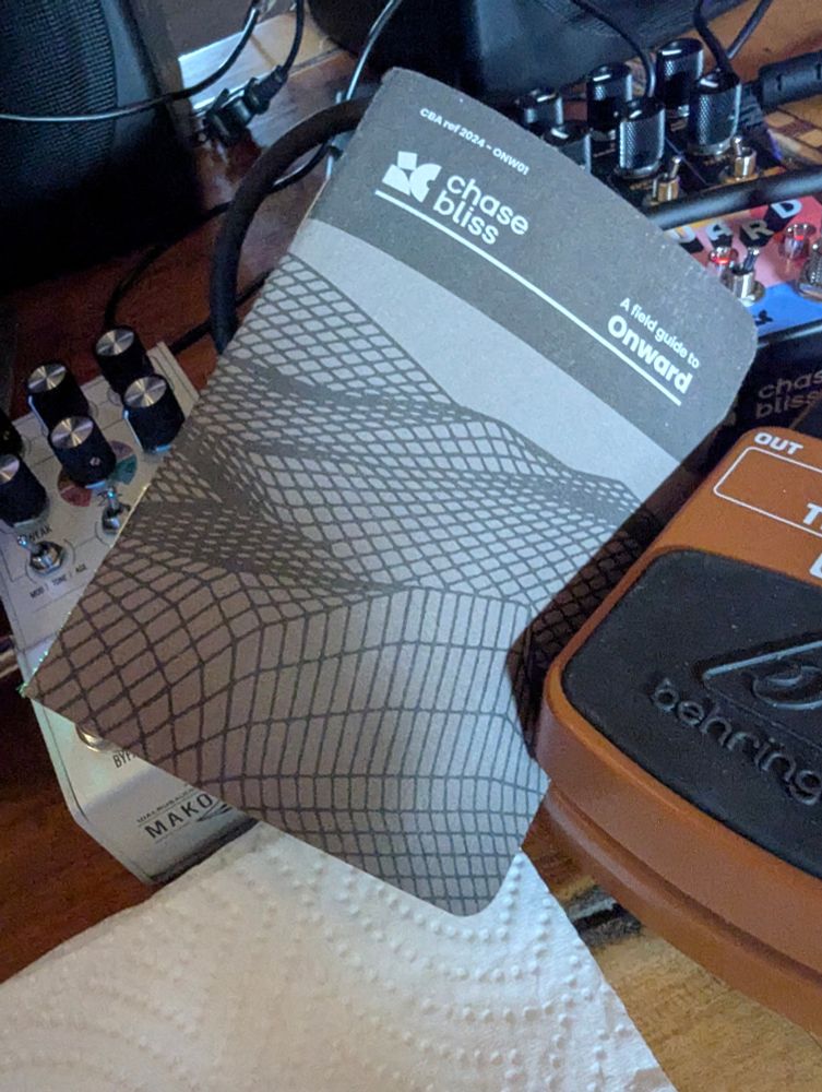 Image dominated by a manual cover for Onward pedal. It hides a D1 and we see the onward peeking out. The Tremolo pedal holds the manual wedged in. Everything sitting on damaged veneer tabltop