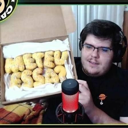 Youtuber Casemiro, com fones de ouvido e em frente a um microfone, mostra uma caixa aberta com salgados em forma de letra, formando a frase "METEU ESSA?".