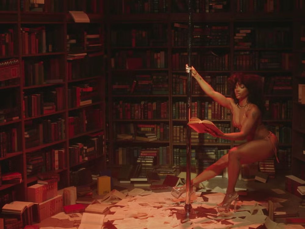 imagem da cantora sza, uma mulher negra belissima, de biquíni, ao dançando num pole dance dentro de uma sala de biblioteca ao mesmo tempo que lê um livro