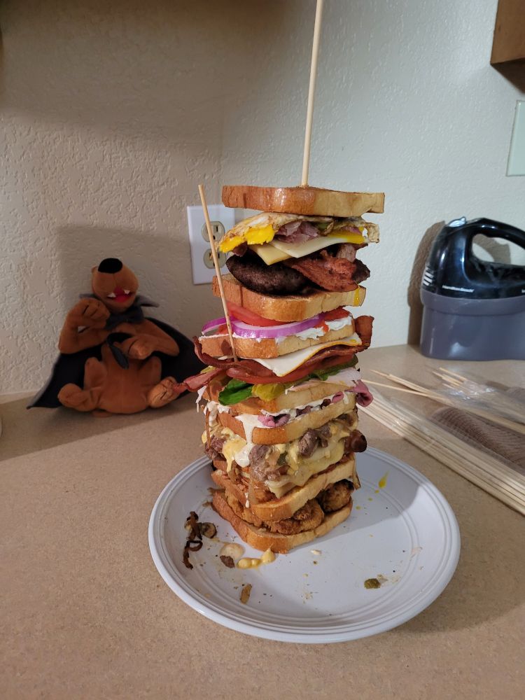 A tower of a sandwich stands on a plate supported by a couple of dowel rods. It stands about 7 or 8 layers of different kinds of sandwich "decks". A stuffed animal Halloween addition Scooby-Doo doll sits in the background 