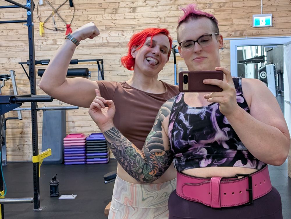A picture of me with a full sleeve tattoo, pink hair, and a pink weightlifting belt on. Eve is behind me with red hair sticking out her tongue and flexing her left arm as I point at it. 