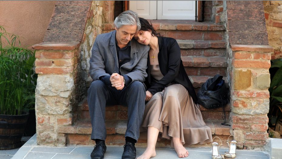 A still from the film Certified Copy: William Shimell and Juliette Binoche sit outside on a brick staircase. He is in a grey suit, she is wearing a taupe satin dress with the black jacket. She is barefoot, her high heels removed slightly off to the side of her feet. She is resting her head on his shoulder. 
