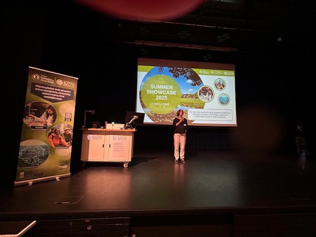 Prof Tina Duren stands on stage in front of a PPT and banner of the Summer Showcase 2025