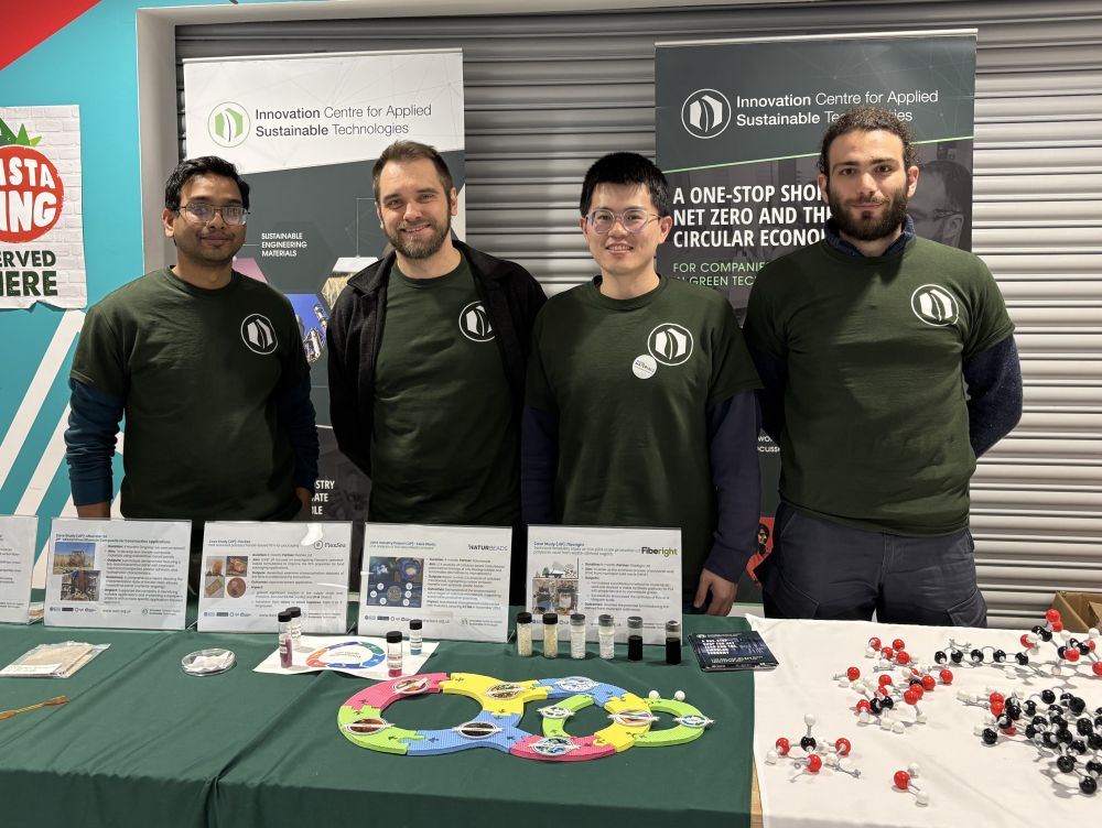 Four members of the iCAST team in dark green branded jumpers stand behind a stall with interactive games and display boards.