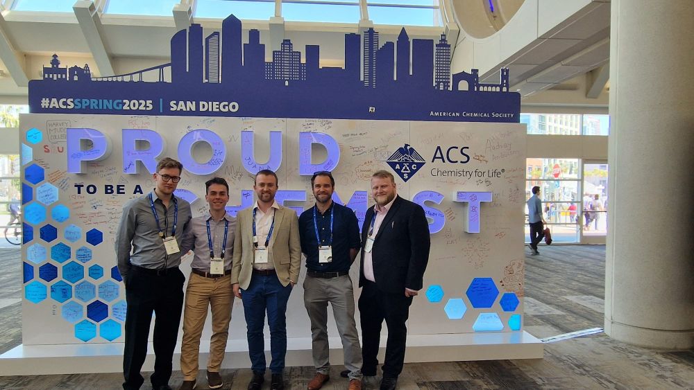 Dominic Savage, Matthew Cullen, Strachan McCormick, Jon Husband and Andrew Kay stand in front of a large poster saying 'Proud to be a chemist'.