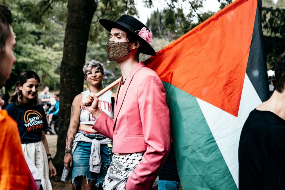 Spice outside in woodland wearing a pink blazer, black hat with pink rose in the brim. Around their waist a Kaffiyeh is tied, & they're holding the flag of Palestine across their back.