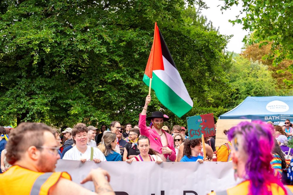 Spice at Bath Pride. In amongst an assembled mass of people, Spice stands in a pink cropped blazer with a gold face mask & black hat, waving the flag of Palestine at the head of the start of a march.