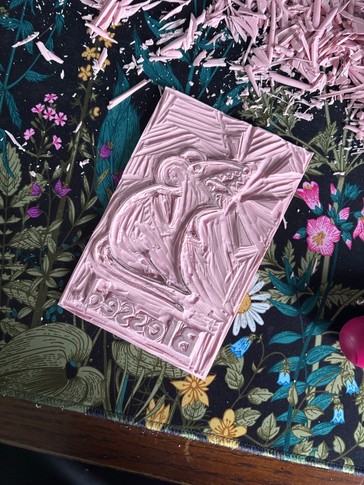 Linocut slab with yelling rat and the word”blessed” beneath it. In the process of cutting away to leave the outline for printing. There’s a pile of cut slivers just above the slab. 