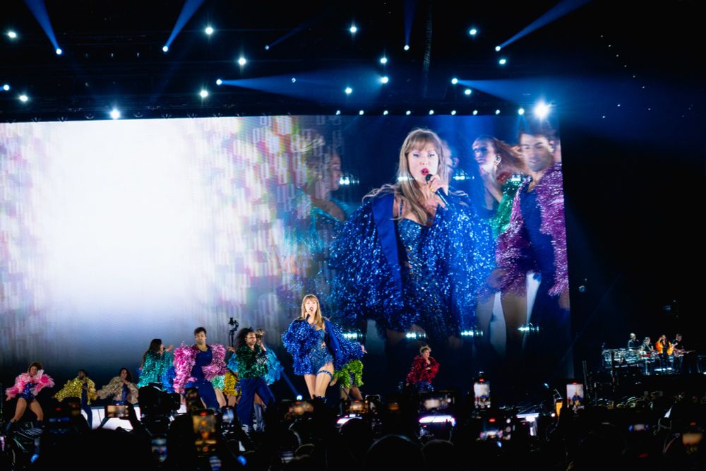 Taylor singing into her mic during Karma - you can see her chevron blue bodysuit cutouts, her shimmery blue jacket, and her garter. Silhouettes of fans' hands in the crowd reaching for taylor are visible at the bottom of the image 