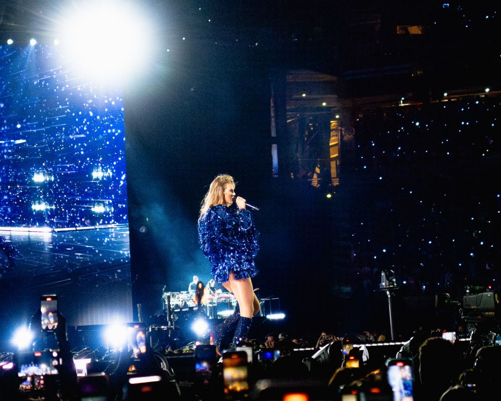 Taylor walking toward the crowd, microphone to her mouth, she is wearing a blue fringe karma jacket and blue knee high glittery boots