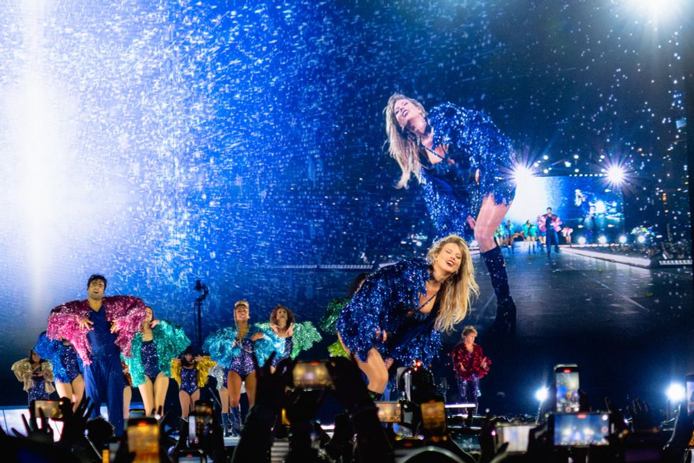 Taylor with her head cocked, hair flipped to the side, her blue karma jacket shimmering, and her dancers behind her