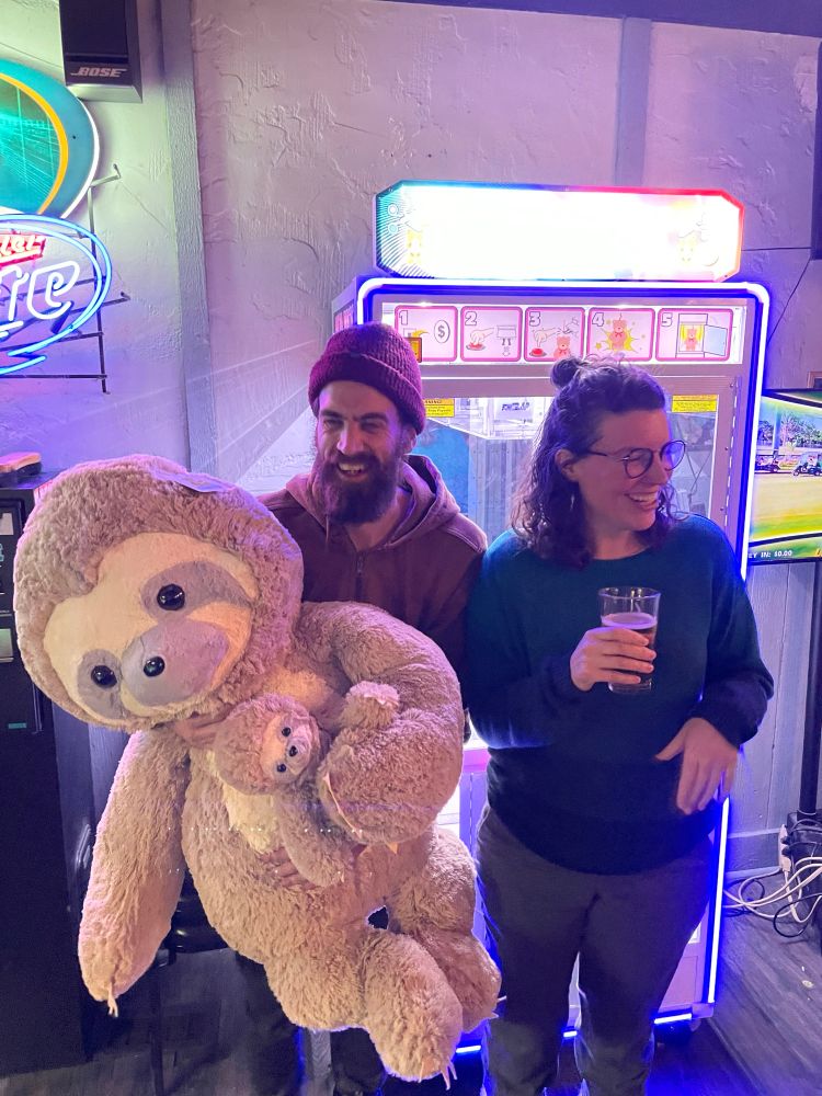 Me holding a giant sloth stuffed animal in front of an arcade game in a bar, with my wife