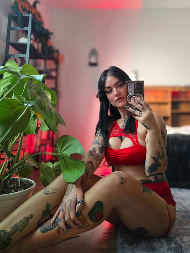 Mirror selfie of a tattooed woman wearing red lingerie seated on the floor by a large houseplant