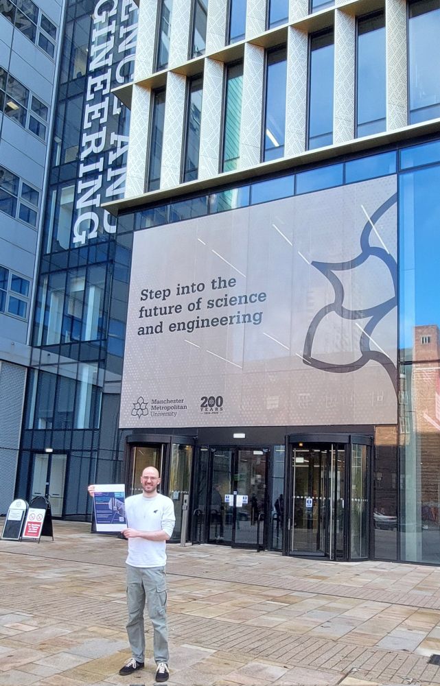 The image shows a man (Kammy Bogue) holding a promotional poster for the Carers In STEMM Survey, in front of a sign for Manchester Metropolitan University. The sign reads "step into the future of science and engineering". 