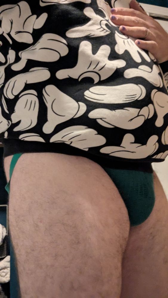 3/4 frontal selfie wearing just a blue and green jockstrap and a sweater with a black background filled with Mickey glove hands in white.  Only the green of the pouch of the jockstrap can be seen in front, and then some of the green leg strap can be seen wrapping around the rear