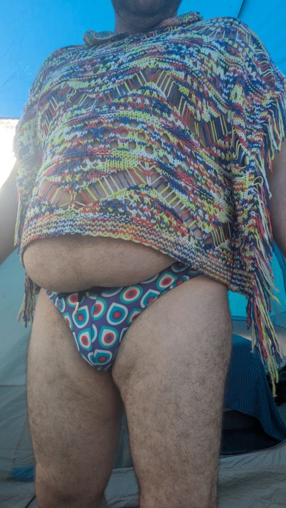 Mostly frontal selfie wearing a blue/red/purple speedo and a blue/orange/yellow/white knitted poncho.  The belly is hanging out the bottom of the poncho and hanging over the waistband of the speedo