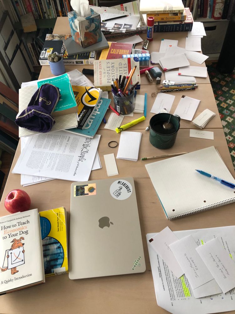 A table covered with study materials: books, flashcards, highlighted notes, pencils, pens, highlighters, laptop as well as otc medications, kleenex, an apple, a mug of tea, a crown royal bag i keep my flashcards bundles in, a small plant (lithops?) a lighter etc….