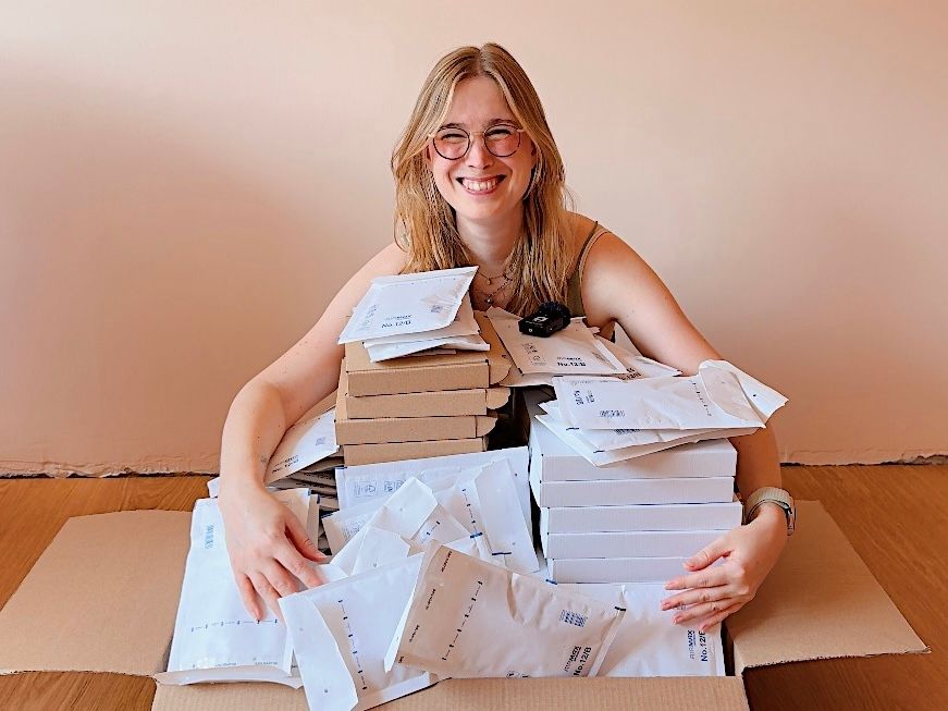 A photo of a smiling blond woman sitting behind a great big stack of boxes and packages with her arms around the pile.