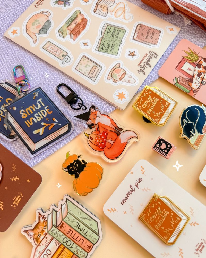 A photo of a flatlay with a variation of cute products on it, such a fox witch keychains, pins of pumpkin cats, tiny books, or books reading 'cozy reader' and stickers, stickersheets and a magnets reading "too many books, too little time". 
