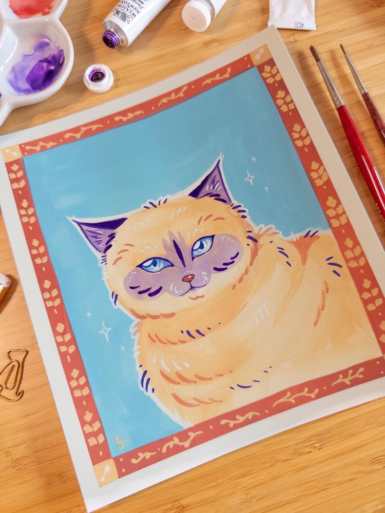 A colorful portrait of a cat made in gouache and posca markers. The cat is beige with purple shadow markings in it's snoot and on it's ears. The drawing is surrounded by traditional art supplies like markers, paint tubes and brushes.