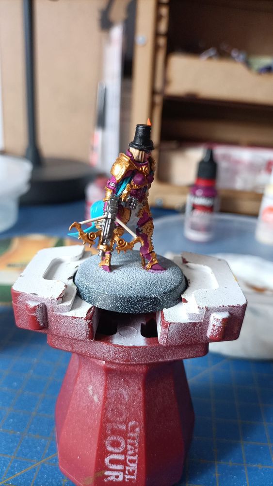 Foto de la miniatura de la inquisidora Greyfax de W40k con los colores base dados vista desde su derecha. Lleva armadura dorada y magenta, sombrero negro con una vela, faldón blanco, una capa turquesa y una ballesta en la mano derecha 