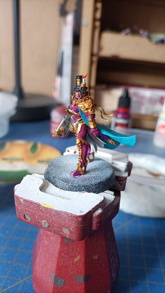 Foto de la miniatura de la inquisidora Greyfax de W40k con los colores base dados vista desde su izquierda. Lleva armadura dorada, magenta, sombrero negro con una vela, una capa turquesa y una ballesta en la mano derecha 