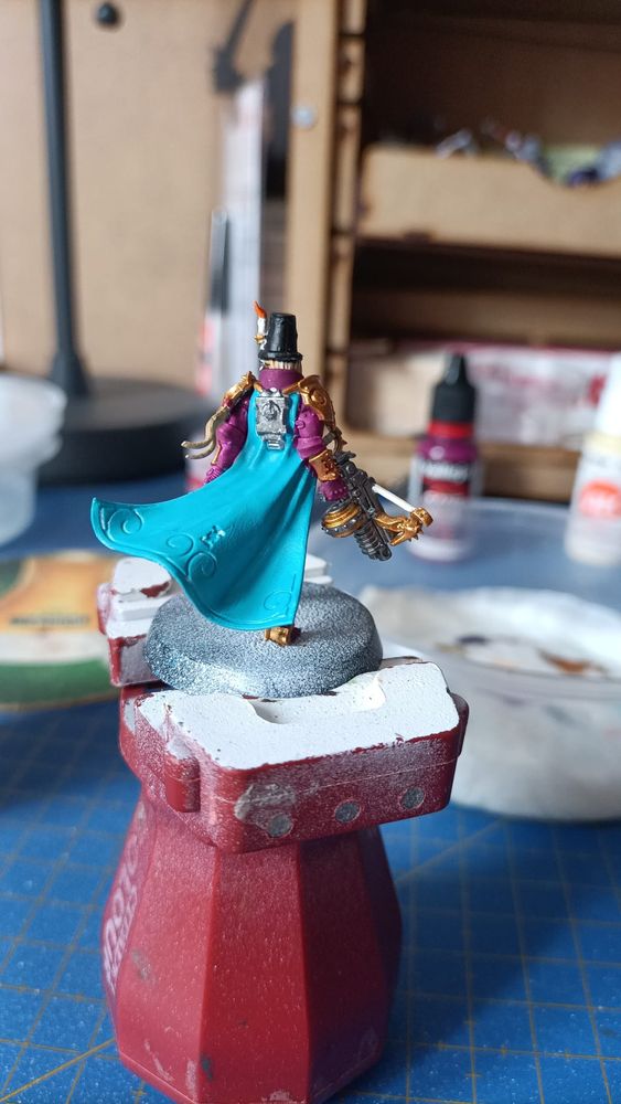 Foto de la miniatura de la inquisidora Greyfax de W40k con los colores base dados vista por detrás. Lleva armadura dorada, magenta, sombrero negro con una vela, una capa turquesa y una ballesta en la mano derecha 