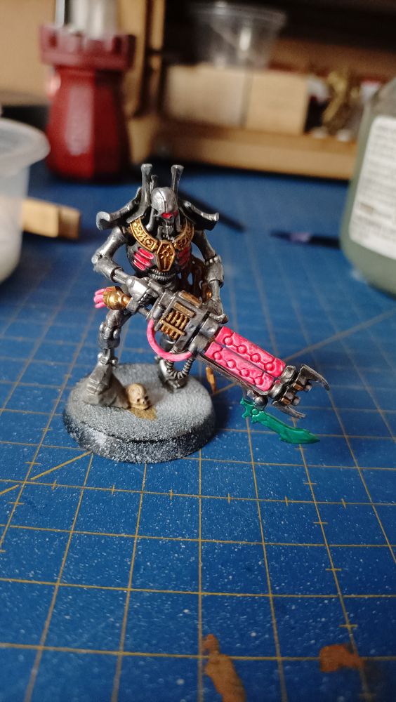 Miniatura de un necrón guardia real de Warhammer 40k en proceso de pintado, principalmente metálico plata y detalles oro, y fluo rojo en el arma, cables, ojos y aberturas intercostales 