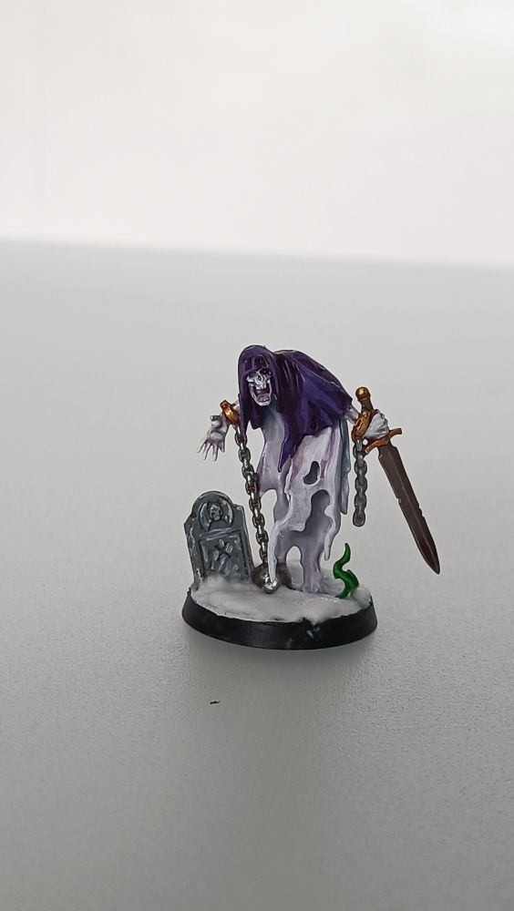 Miniatura de un fantasma de Warhammer Age of Sigmar con harapos blancos y capucha morada, que arrastra cadenas y sujeta una espada, y una lápida de piedra a su lado