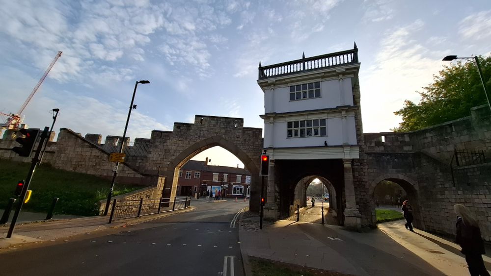 Medieval wall gate in York, 2 arches one with building on top of it, blue sky with light clouds and rays of sun