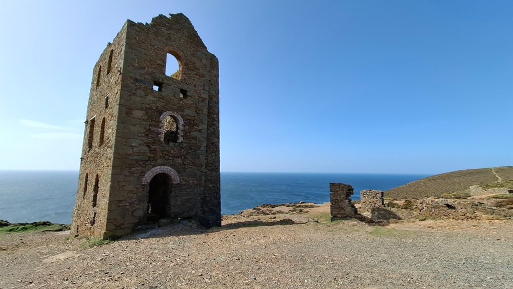 Abandoned mining building, on cliff a stone 3 storey building with many windows. Remainds of other buildings visable and the sea behind, blue sky